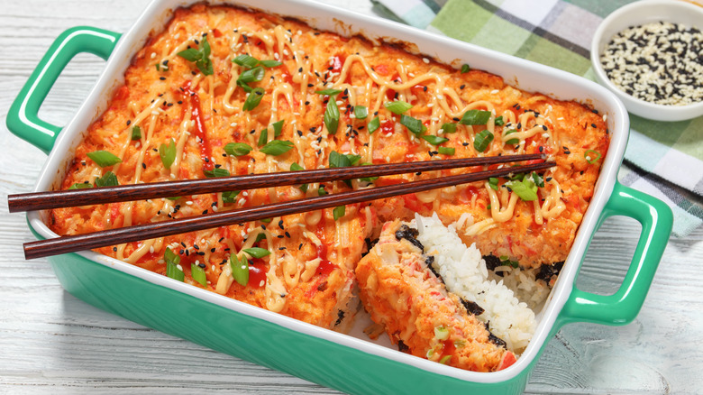 A green baking tray filled with sushi bake drizzled with spicy mayo and green onions with chopsticks on top