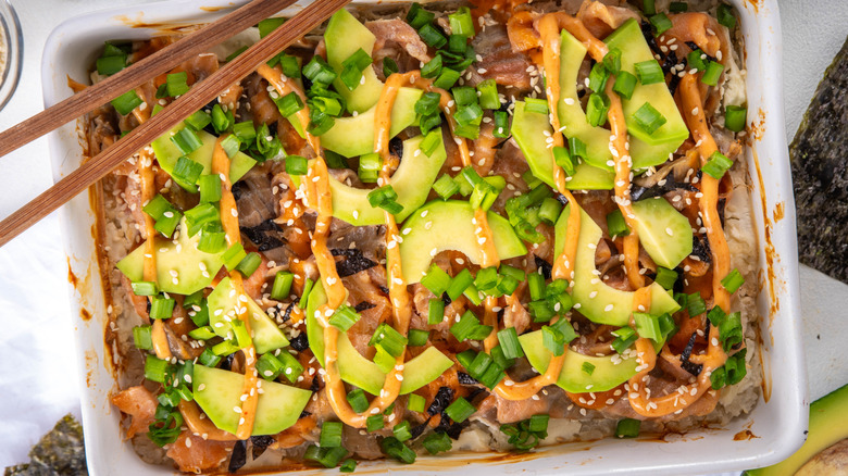 A baking tray of sushi bake garnished with fresh avocado, sesame seeds, and green onions