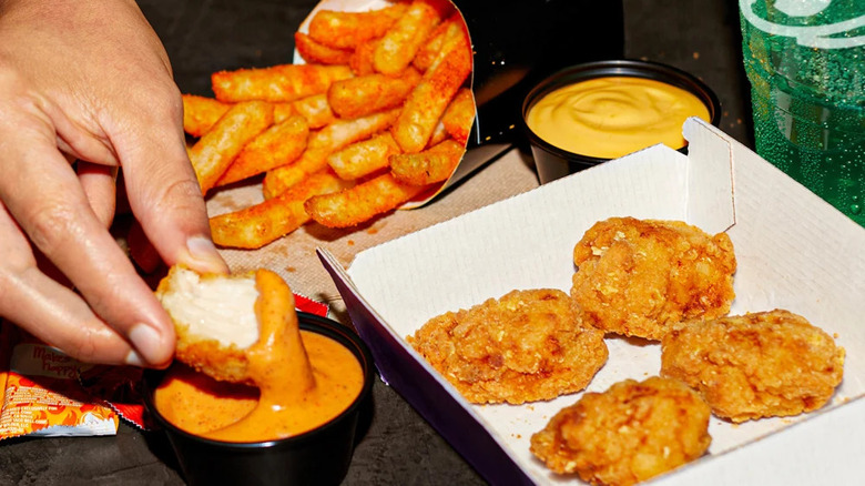 A hand dipping a Taco Bell crispy chicken nugget in orange sauce with other nuggets, fries, and cheese sauce beside it.