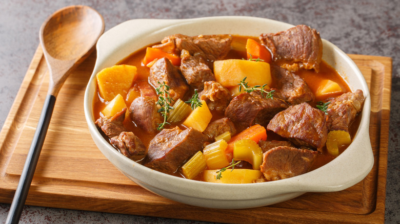 A bowl of beef stew on a wooden board with a spoon