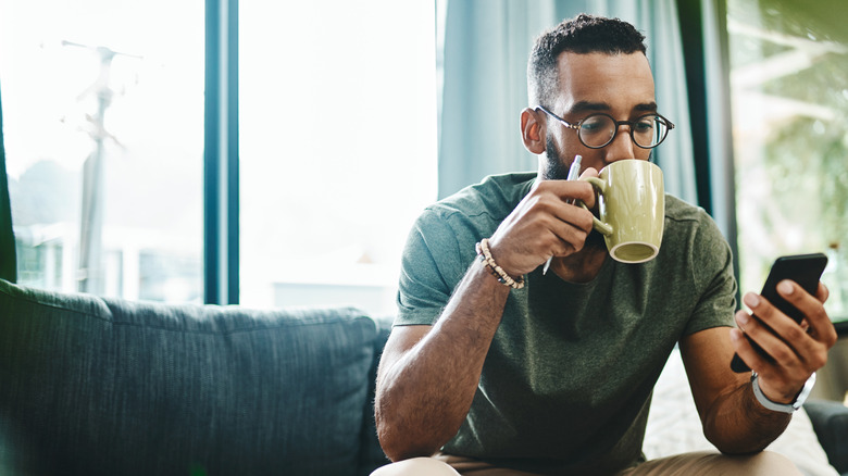 Person drinking coffee on couch while looking at phone