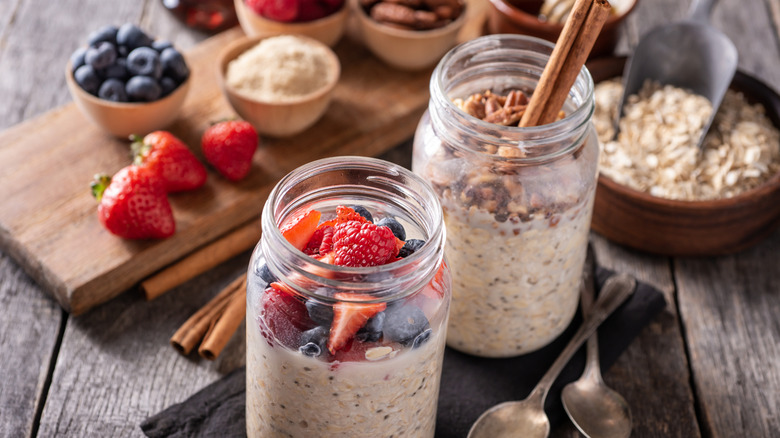 two mason jars of overnight oats topped with fresh berries and nuts surrounded by assorted berries, oats, and cinnamon sticks