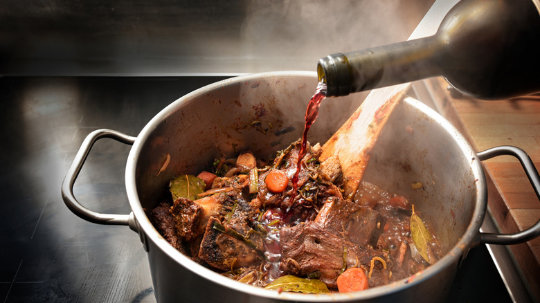 A bottle of red wine being poured into a pot of meat and vegetables