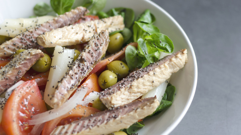 Slices of seasoned pinkish fish sits atop a salad with tomatoes, olives, and spinach