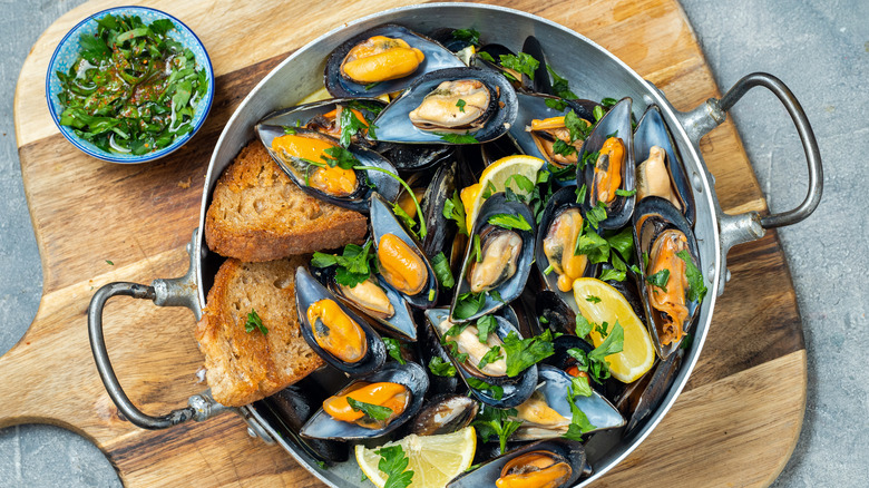 A metal pot filled with orange muscles, sprinkled with greens, slices of lemon and toasted bread. All atop a wooden cutting board