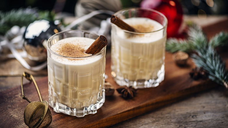 Two glasses of eggnog with nutmeg and cinnamon sticks on wood board