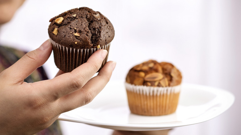 hand holding a dark brown muffin with a lighter muffin on a plate in the background