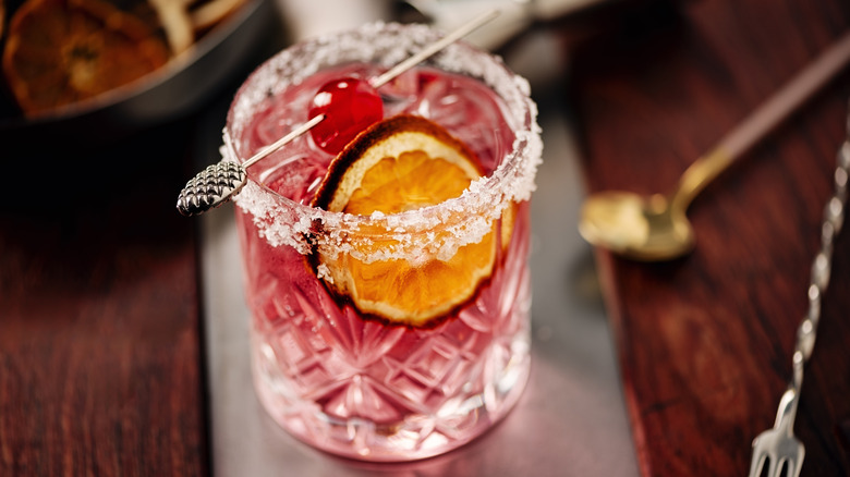 A pink mocktail in a sugar rimmed rocks glass with a maraschino cherry garnish