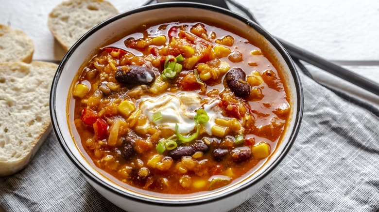 Bowl of chili with sour cream and scallions with bread in background.