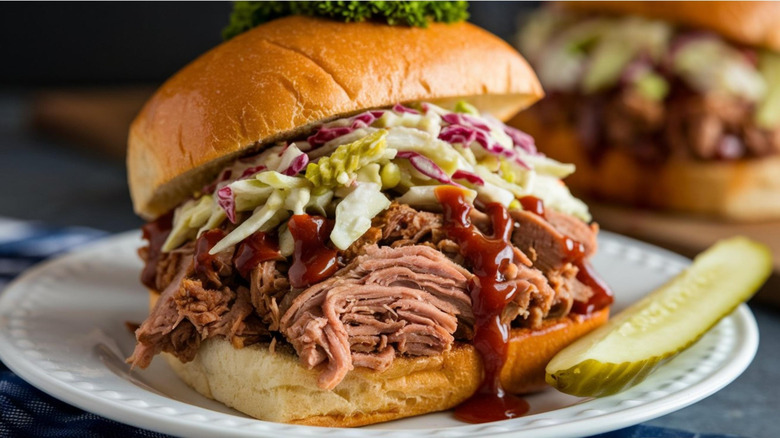 A BBQ sandwich with coleslaw on a white plate with a pickle on the side.