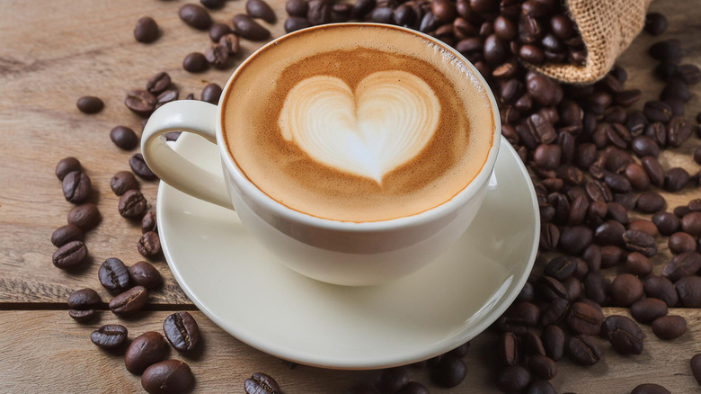 Latte with a heart made of foam in a white coffee cup on a saucer sitting on a wooden surface and surrounded by spilled coffee beans from a burlap bag.