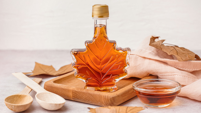 Clear maple syrup bottle in the shape of a maple leaf on a wooden cutting board surrounded by dried maple leaves and wooden spoons.