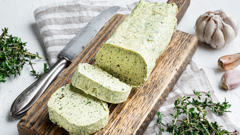 A log of a green compound butter with herbs and garlic on a wooden board with a butter knife on the side