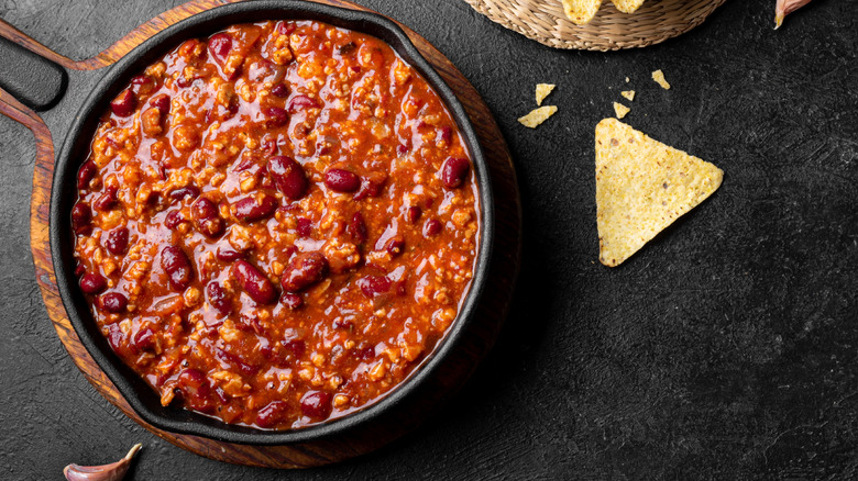 A cast-iron pan full of chili with a single tortilla chip beside it on a black surface.