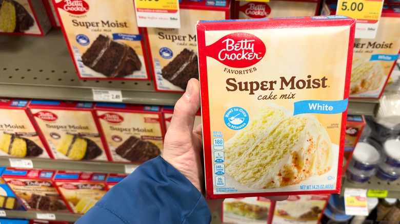 Person holds a box of super moist white cake mix in a grocery store aisle, in front of shelves of other boxed cake mix.