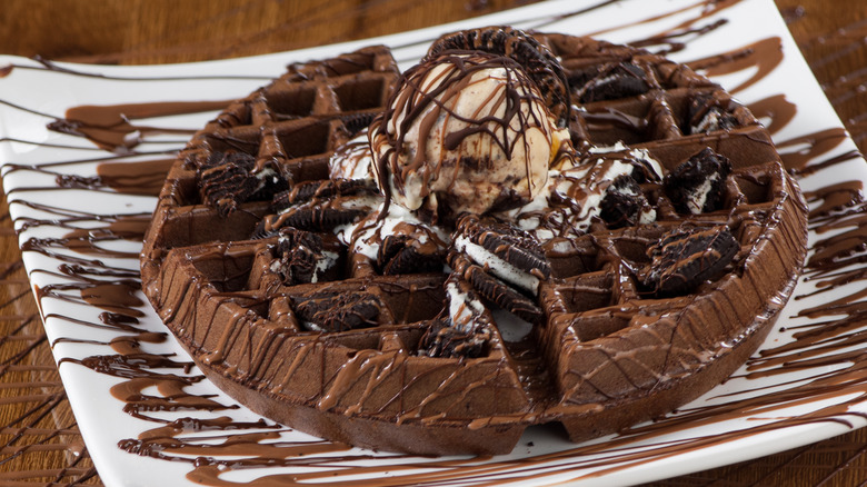 A chocolate waffle topped with oreos and ice cream sits on a white plate, drizzled with caramel and chocolate