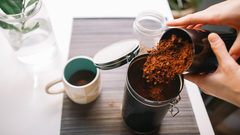 Person dumping ground coffee into a container with a cup of coffee nearby