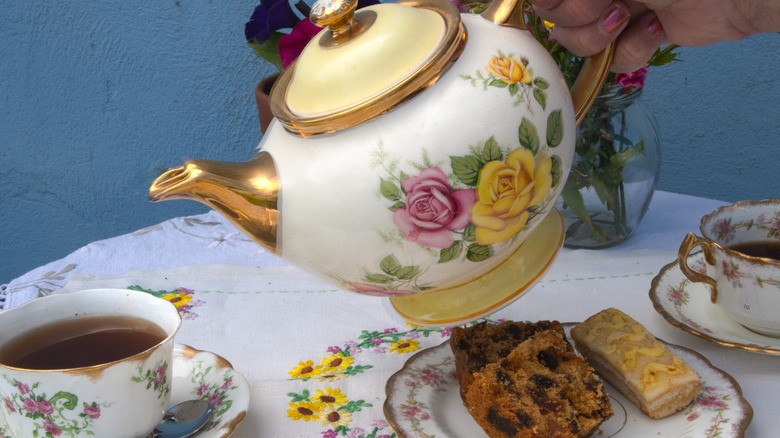 hand holding a floral-patterned teapot over a floral-patterned cup of tea and a floral-patterned plate with raisin bread and a small slice of cake