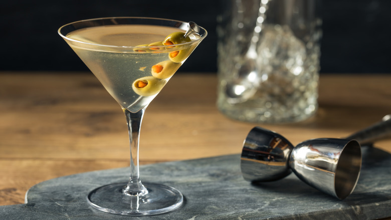 A dirty martini with olives beside a measuring shot glass