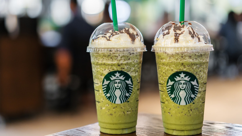 Two matcha lattes in Starbucks takeaway cups topped with whipped cream and green straws