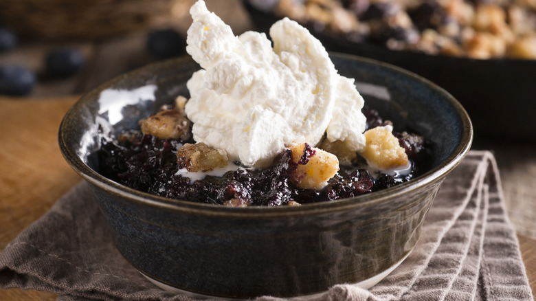 Blueberry cobbler with whipped cream on top in a dark bowl on a cloth napkin.