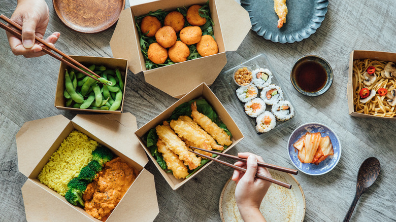 A variety of takeout foods in cardboard tubs being eaten with chopsticks