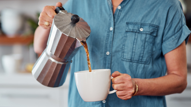 A woman pours coffee from a Moka pot into a mug