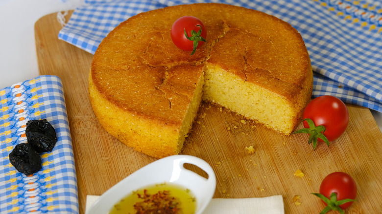 A round cornbread loaf on a wooden chopping board with fresh tomatoes and a slice cut out