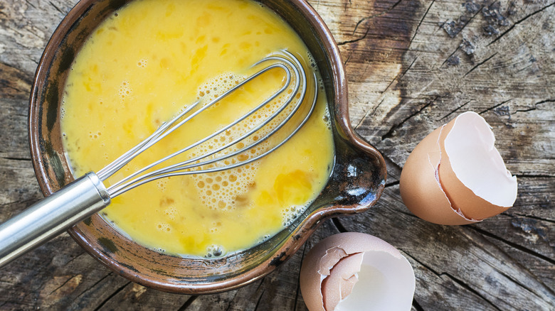 Eggs being whisked in a ceramic bowl on a wooden surface with two broken egg shells next to the bowl