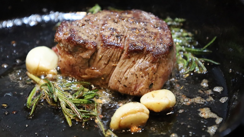 A juicy filet mignon cooks in a cast iron pan with garlic and rosemary