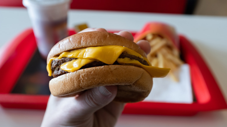 someone holding a nondescript fast food burger with fries and drink in background