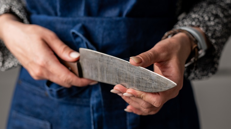 Woman Checks Knife Sharpness In Blue Kitchen Apron, Close-Up Of Hand In Focus