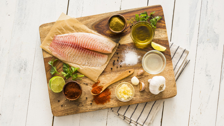 A tilapia filet on a cutting board, surrounded by small bowls of seasonings.