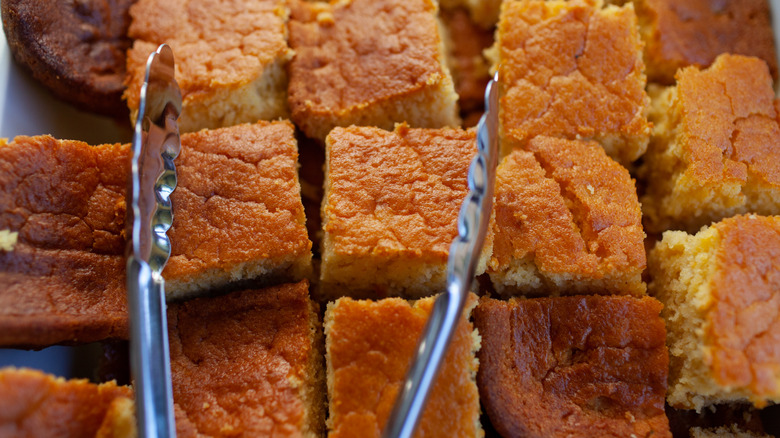 Freshly made squares of cornbread sit on a baking sheet with a pair of tongs.