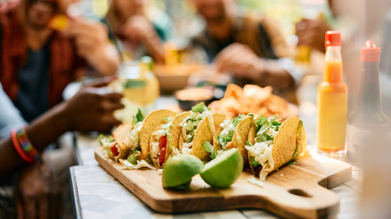 Close-up of tacos on a wooden cutting board with blurred friends in the background