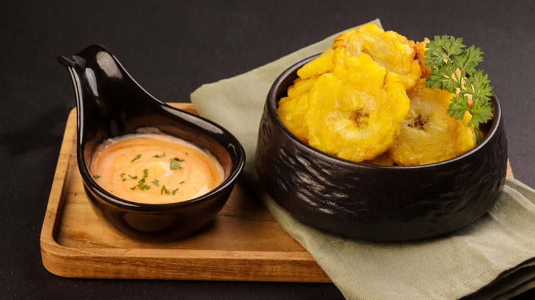 wooden tray holding bowl of fresh, crispy tostones with herb garnish beside small bowl of creamy sauce for dipping