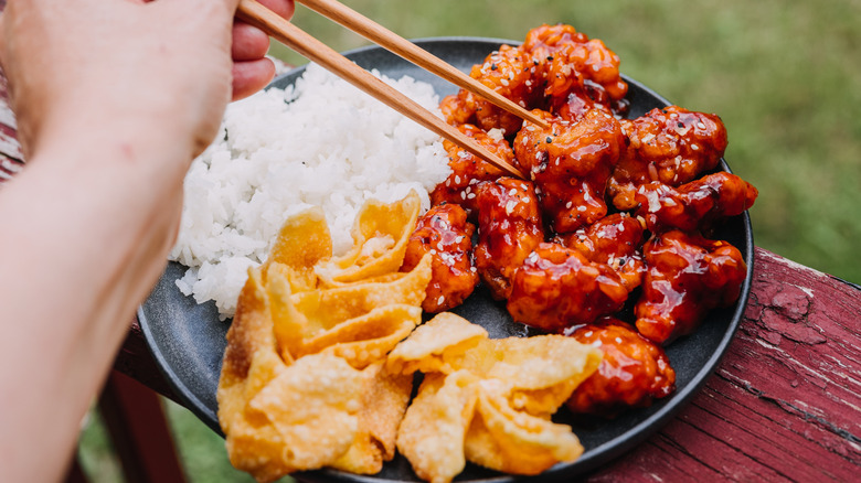 Person using chopsticks to pick up Chinese food with white rice and crab rangoon on the side