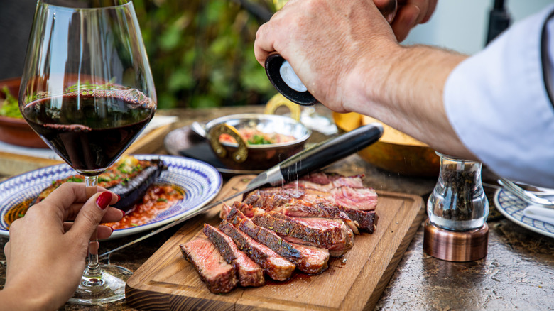 A person seasoning sliced steak on a wooden board at a dining table