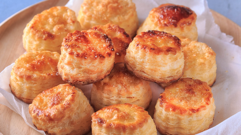 A pile of flaky, crispy buttermilk biscuits on a parchment paper-covered wooded surface.