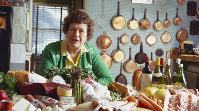 Julia Child in her kitchen with produce in front of her, and copper pans on the wall