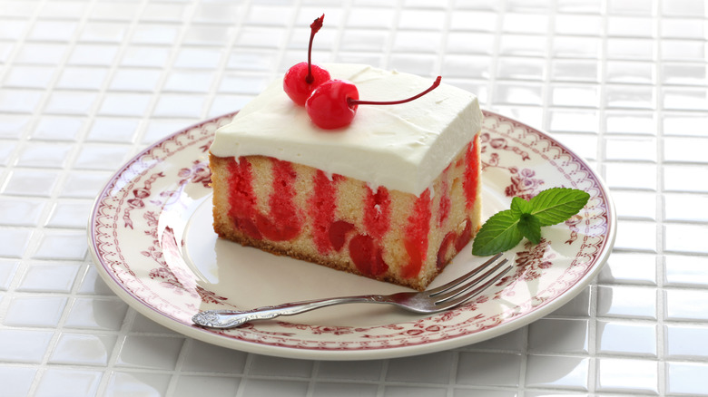 Vanilla cherry poke cake with cream cheese frosting and two maraschino cherries on a white and maroon plate with a dessert fork and mint leaf