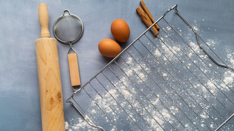 A cooling rack on a grey counter surrounded by baking tools, flour, and eggs