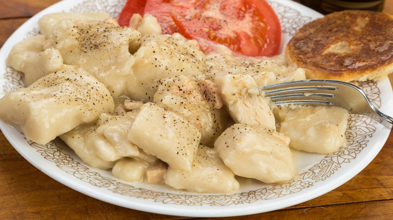 Chicken and dumplings served on a plate with a biscuit and tomato slices