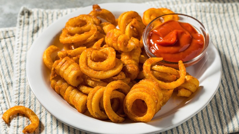 A plate of curly fries on a linen towel, with a small glass bowl of ketchup for dipping.