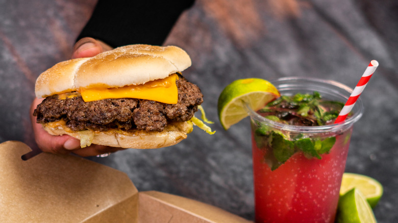 A hand holds a smash burger next to a cocktail