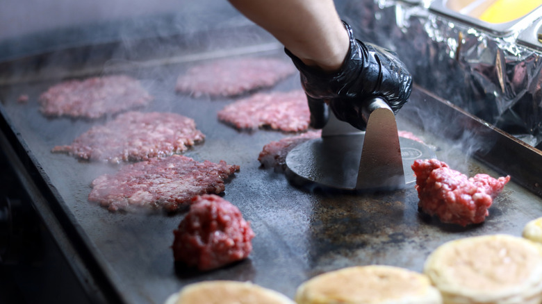 A cook presses down burger meat on a grill for a smash burgers