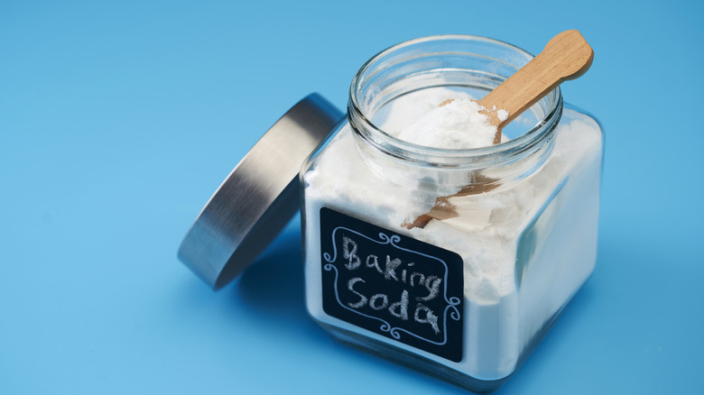 Cube glass jar of baking soda with a wooden spoon on a blue background.