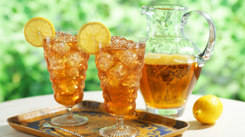 Two crystal cups full of iced tea sitting on a blue and mustard tray with ice and a lemon slice for decoration. Behind the tray the pitcher, half full sits next to a lemon on a white surface.