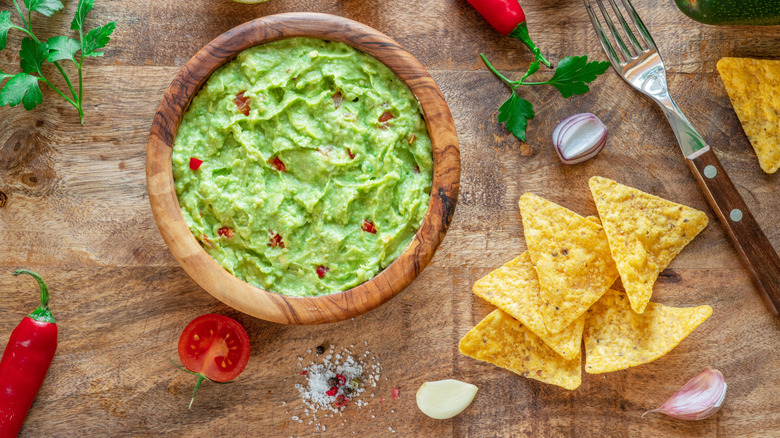 A bowl of guacamole surrounded by red peppers, lime, tortilla chips, tomato, a fork, and garlic cloves.