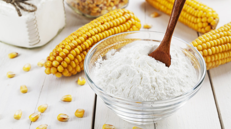 A glass bowl of cornstarch on a wooden surface with fresh corn cobs